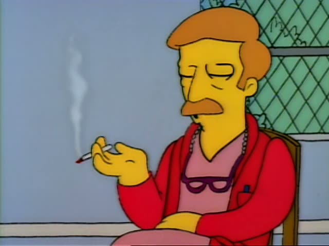 A seated, mustached man with orange-red hair, a red jacket over a pink top, a beaded necklace and purple glasses smokes…