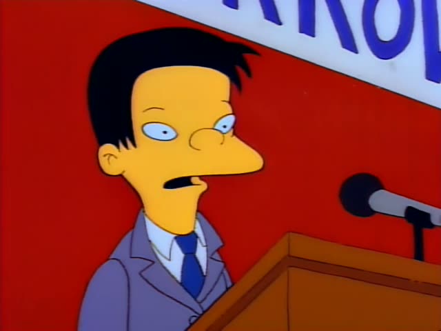 A thin, dark-haired man in glasses, a grey suit and a blue tie stands behind a wooden podium with a microphone on the right…