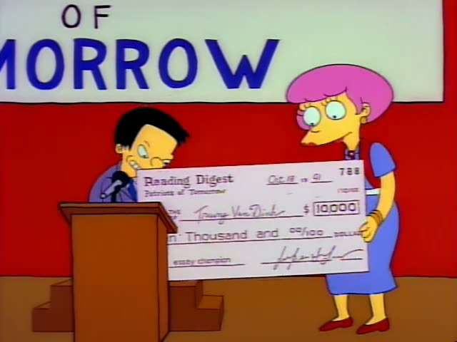 A thin, dark-haired man in glasses and a purple shirt stands behind a wooden podium with a microphone, while a woman…