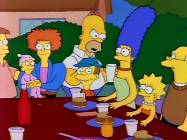 Homer, Marge, Lisa, Bart and Maggie sit with a crowd of relatives and neighbors at a long backyard table crowded…