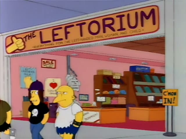 Homer stands outside The Leftorium beside a woman in a purple beanie, black skull shirt, and long brown hair, and a woman…