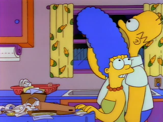 Marge and Homer stand at the kitchen counter beside a cluttered pile of papers, a wooden board and a basket of dishes