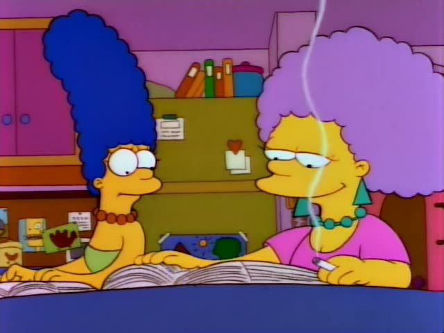 Marge Simpson and her twin sister Selma sit at a table reading a large open magazine in a living room with a green cabinet…