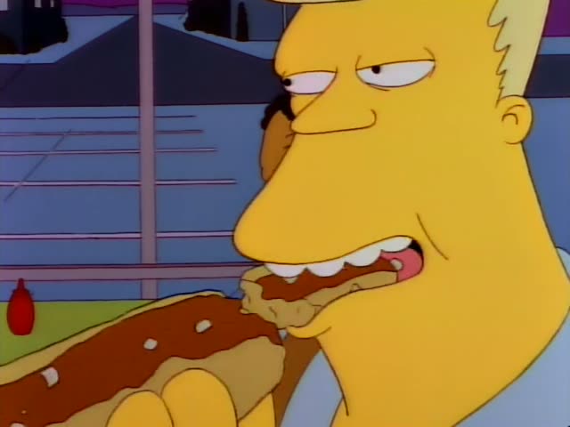 A heavy, light-haired man in a light blue shirt with a shoulder holster bites into a chili-topped hot dog at a green…