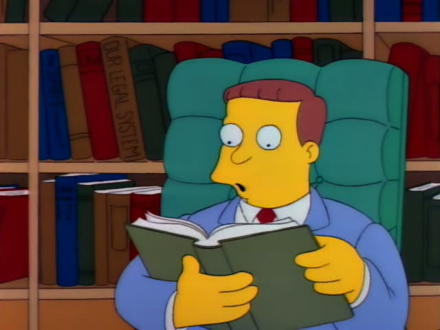 A yellow-skinned cartoon man with reddish-brown hair, a blue suit and red tie sits in a green tufted armchair in front…
