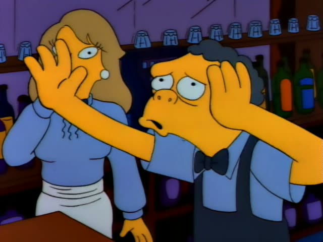 Moe stands beside a woman in a blue sweater and white apron behind a bar lined with bottles, both holding their faces…