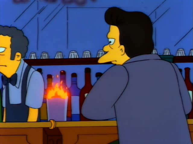 A dark-haired man in a grey coat stands at the bar, watching a glass with flames rising from it, then lifts the flaming…