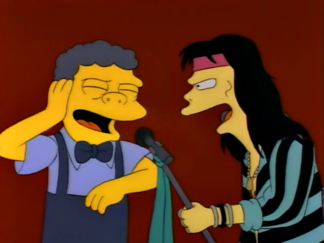 Moe sings into a microphone with his hand cupped to his ear while a long-haired singer in a striped shirt, red headband…