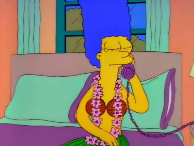 Marge Simpson sits up in bed wearing a grass skirt, a coconut-shell bra and a pink flower lei while holding a purple…
