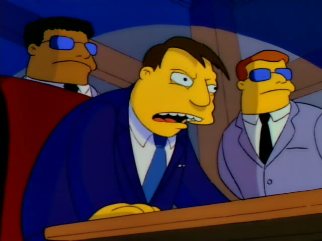 Seated behind a wooden bench in a wood-paneled room, a heavy-set man in a blue suit and tie, with dark hair, grits his teeth…