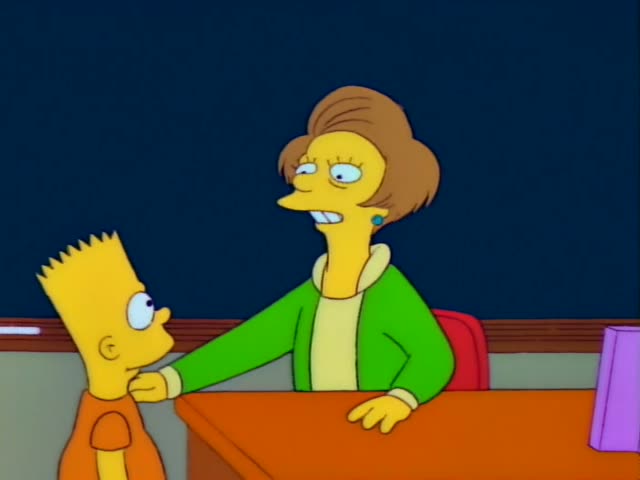 Bart stands at the left beside an orange desk, facing Mrs. Krabappel, his teacher, who has a tall brown hairdo and wears…