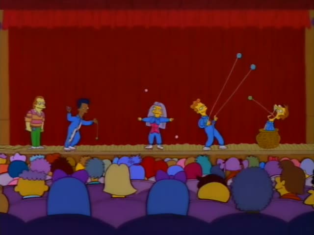 Lisa sits in the audience beside Marge, whose tall blue beehive stands out, as the family and other spectators watch a stage…