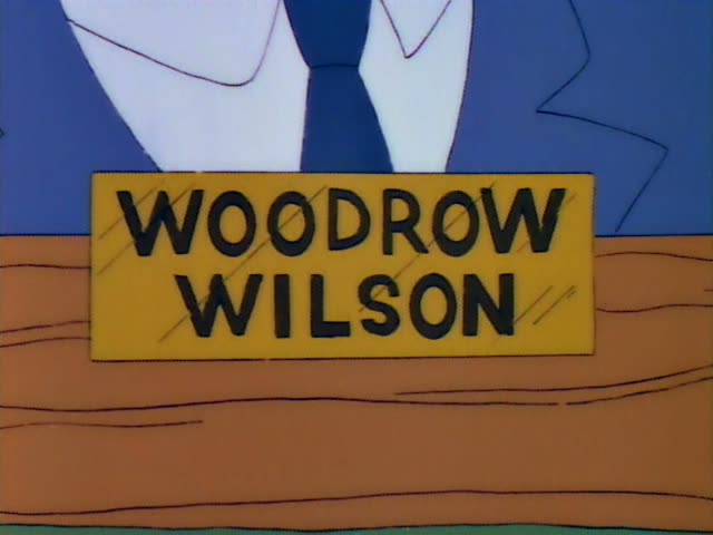 No regular characters are visible; the moment shows a yellow nameplate reading WOODROW WILSON on an orange desk in front…