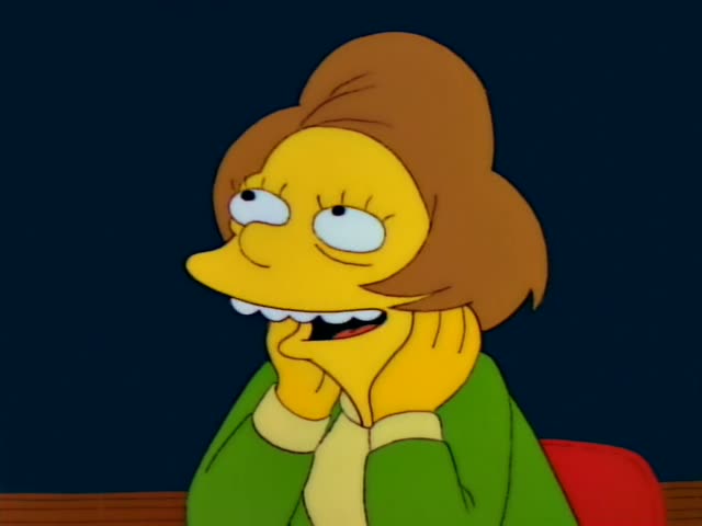 Edna Krabappel sits at a desk with a red chair, first resting her chin on her hands and grinning, then gesturing…