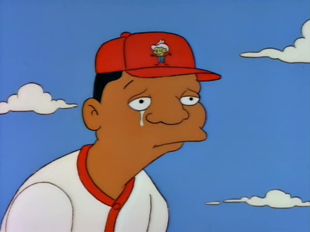 A tearful Black man in a red cap and white baseball jersey with red trim is shown close up, and the scene then opens…