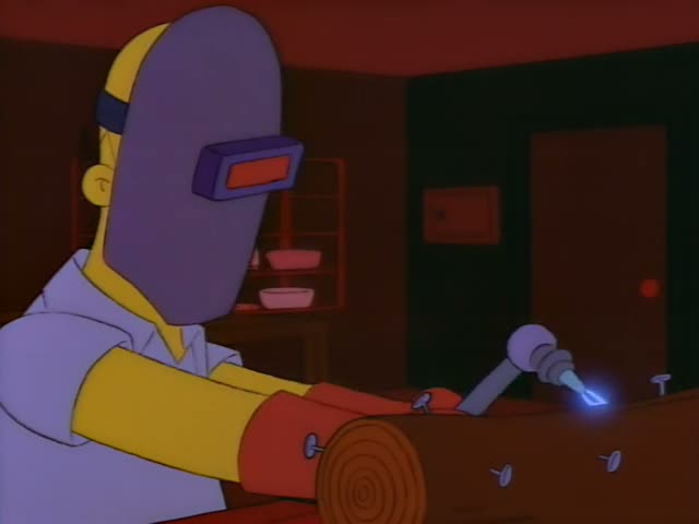 Homer works at a bench in a dim room, sparks flying from a metal piece he is welding onto a wooden log