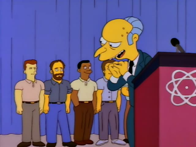 Mr. Burns stands behind a red podium marked with an atom logo, first rubbing his hands together and then extending an arm…