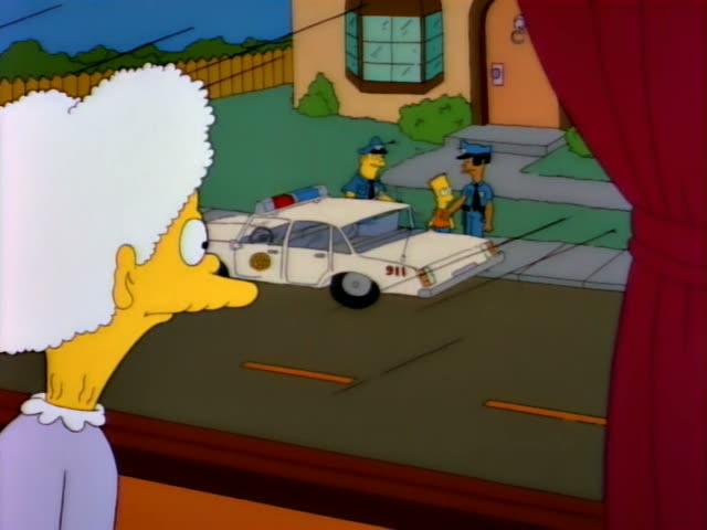Bart Simpson stands beside a white police car on a suburban street while a costumed officer and a grey-haired woman…