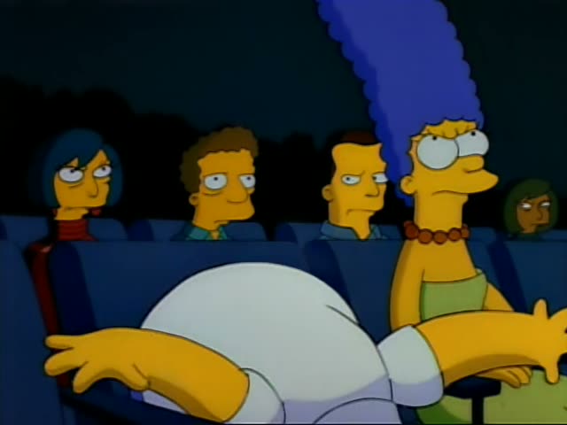 Homer Simpson sits in a dark movie theater row beside Marge, whose tall blue beehive and green skirt are visible…