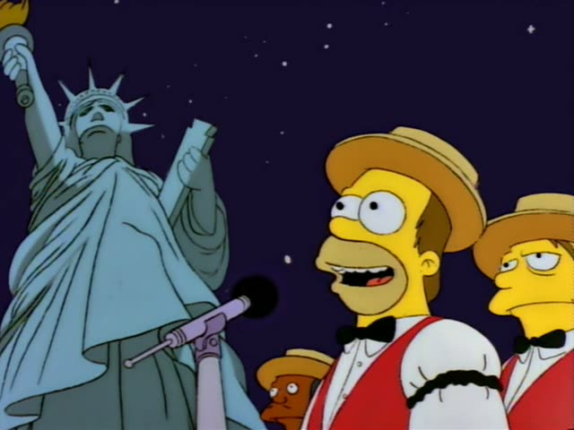 Homer Simpson stands at a microphone beside the Statue of Liberty against a night sky with stars