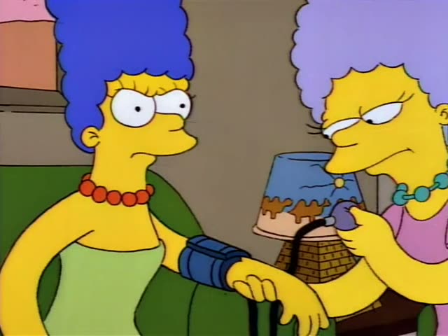 Marge Simpson sits on a green couch with a blood pressure cuff on her arm while her twin sisters, Patty and Selma…