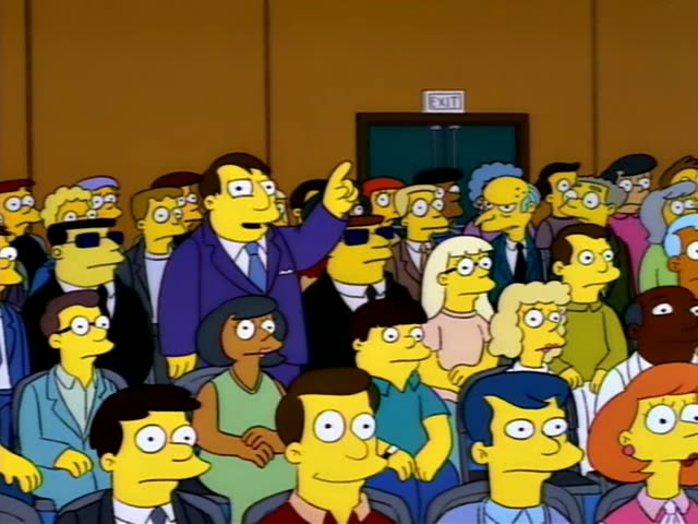 Troy McClure stands in a crowded auditorium beside a row of seated and standing townspeople, many in sunglasses…