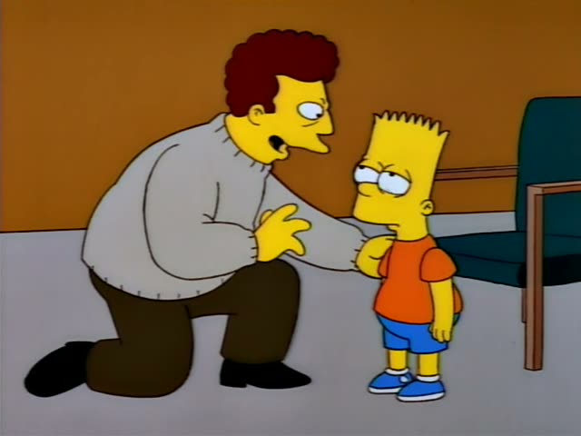 Bart stands in a room with an empty green chair at right while a man with curly reddish-brown hair, a grey sweater and brown…