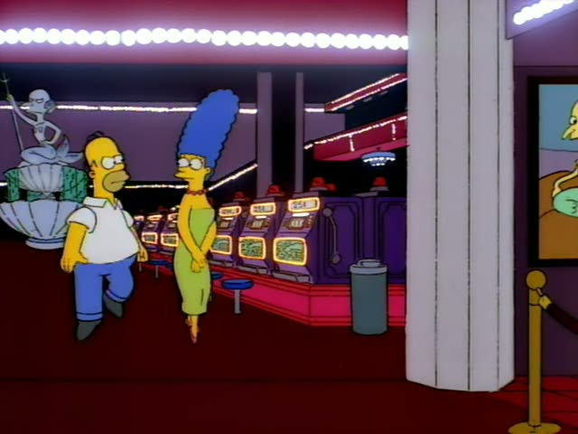 Homer and Marge stand in a casino lobby beside rows of lit slot machines, with a fountain holding a statue and a gold velvet…