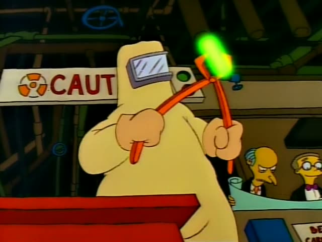 Homer stands at a control counter beneath a CAUTION sign, holding orange tongs that grip a glowing green capsule…