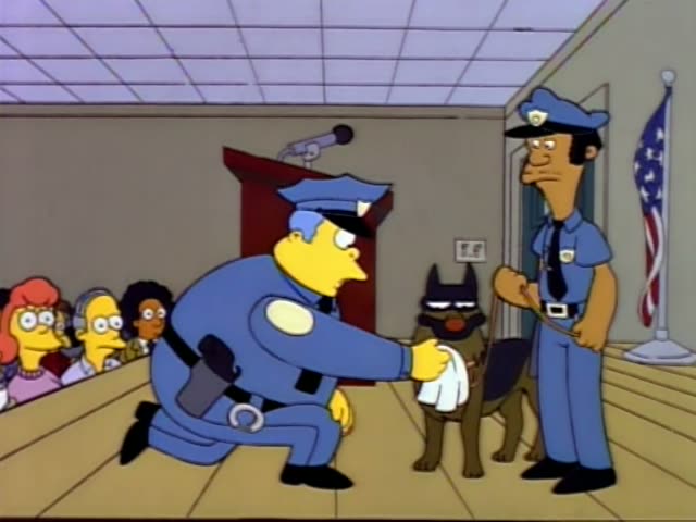 Chief Wiggum kneels on a courtroom floor offering a white cloth to a black-and-tan police dog, while a mustached officer…