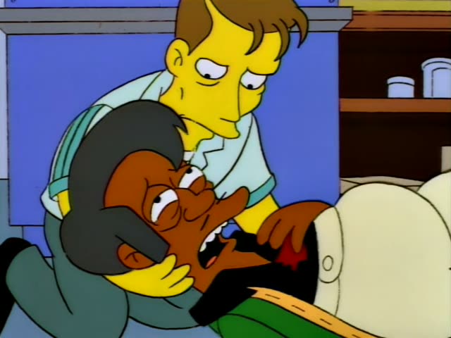 Apu lies in a bed with a red stain on his chest while a brown-haired man in a pale blue shirt leans over him, supporting…