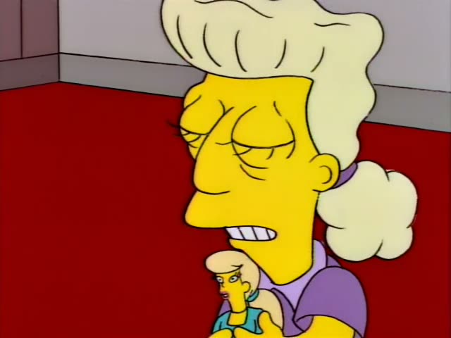 A tall-beehived woman in a purple top grimaces and rolls her eyes beside a young blonde woman in a teal top on a red carpet…