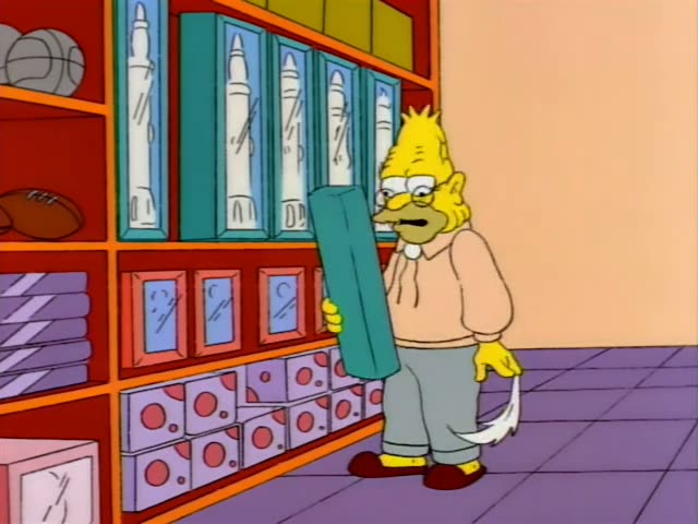 Grampa stands in a toy store among shelves of boxed toys, first holding up a teal box and then a blue and red toy rocket