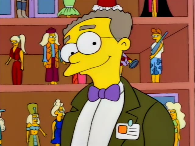 A one-off character, a grey-haired man with glasses, a purple bow tie, a green jacket and a name badge on his lapel, stands…