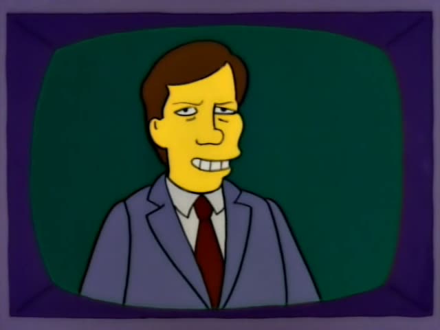 Homer sits on the living room couch aiming a remote at the television, where a brown-haired man in a suit and red tie…