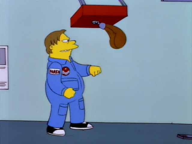 A short-haired man in a blue NASA jumpsuit, who is not one of the regulars, stands in a gym-like room under a red-capped…
