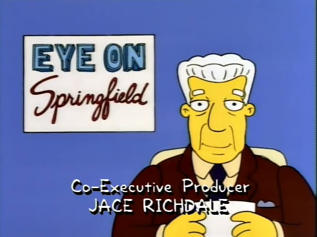 Kent Brockman sits at a desk in front of an Eye on Springfield sign, holding a sheet of paper while on-screen captions…