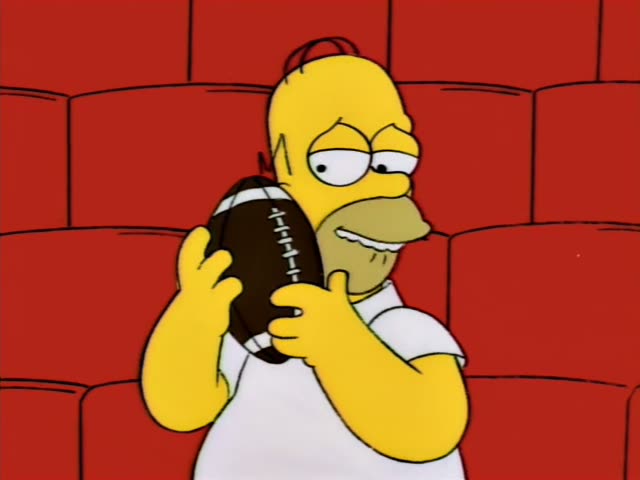 Homer Simpson stands against a red brick-patterned backdrop clutching a brown football with white laces, first gripping it…