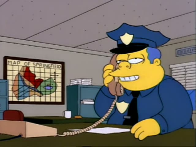 Chief Wiggum sits at a desk in the police station, holding a beige telephone to his ear and talking with an irritated…
