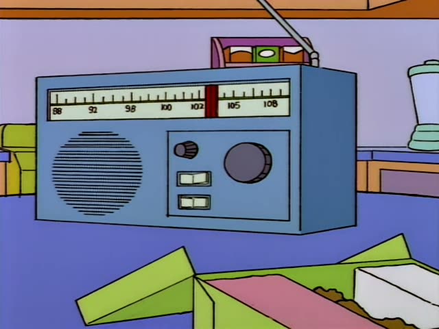 Homer stands at a kitchen table while Bart sits and lifts a small fork toward a plate beside a green box