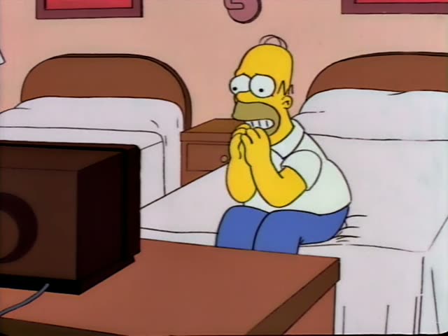 Homer sits on the edge of a bed in a bedroom, facing a dark television on a wooden table in the foreground