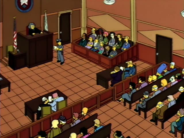 A grey-haired woman judge in a black robe and glasses sits at the bench under a state seal and beside a flag…