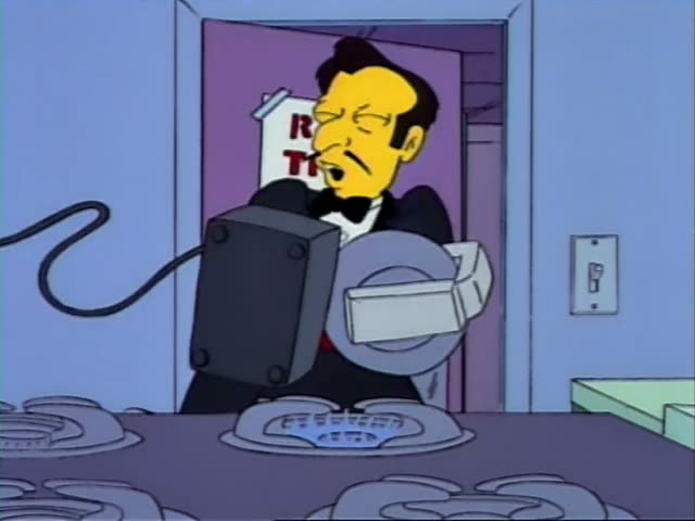 A thin man in a black tuxedo with a bow tie and slicked-back dark hair stands in a kitchen-like room beside a purple door…