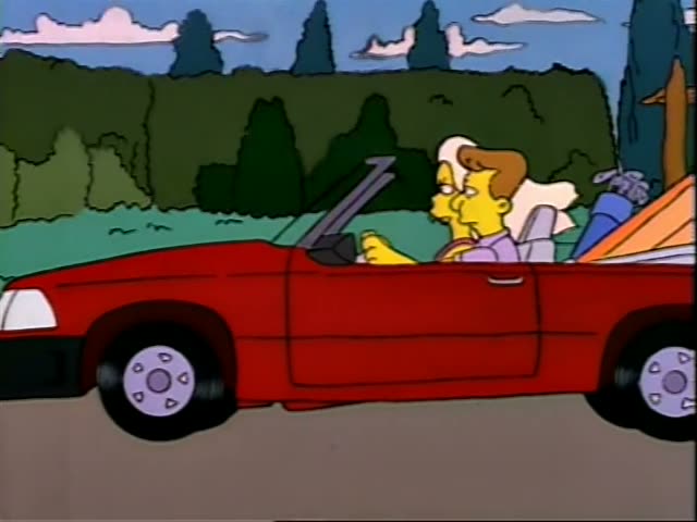 A woman with short brown hair in a purple top drives a red convertible along a road, with a pile of bags in the back seat