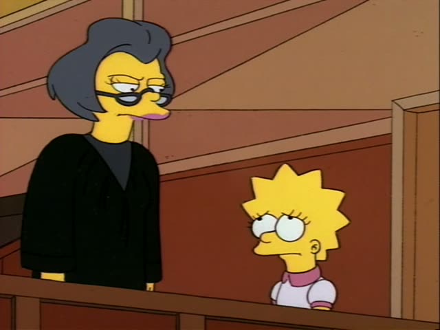 Lisa Simpson stands beside a stern woman with a tall dark bluish-grey bob and glasses, dressed in a black robe, at a wooden…