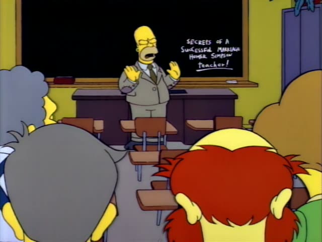 Homer Simpson stands at the front of a classroom in a tan suit, gesturing with both hands before a chalkboard…