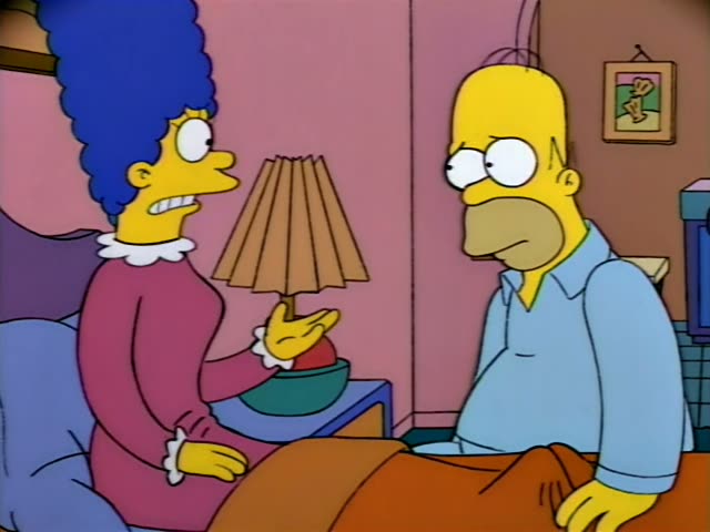 Marge Simpson sits up in bed beside a lamp on a nightstand, while Homer Simpson stands at the foot of the bed on the right