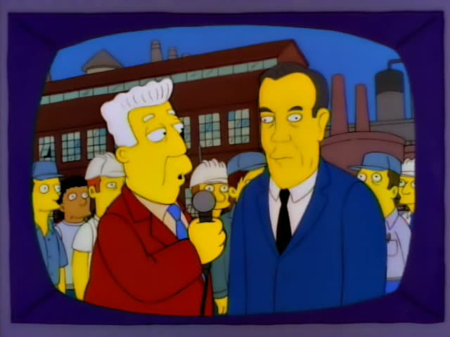 A tall, dark-haired man in a blue suit and black tie stands outdoors before a brick factory with smokestacks…