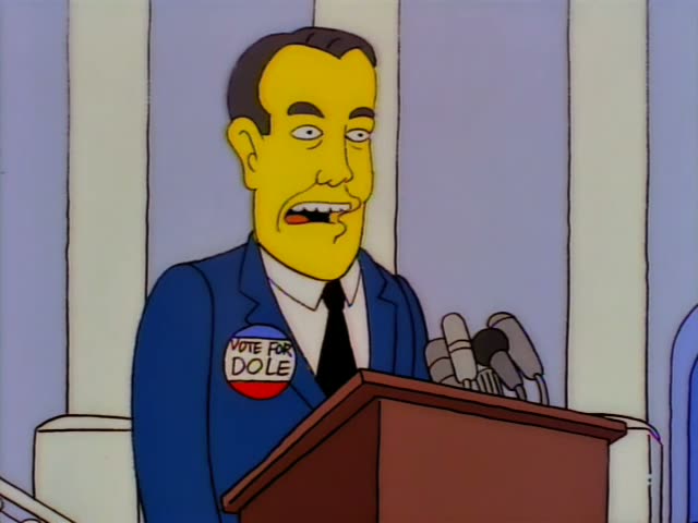 A dark-haired, stout man in a blue suit with a "Vote for Dole" button stands at a wooden podium with a cluster…