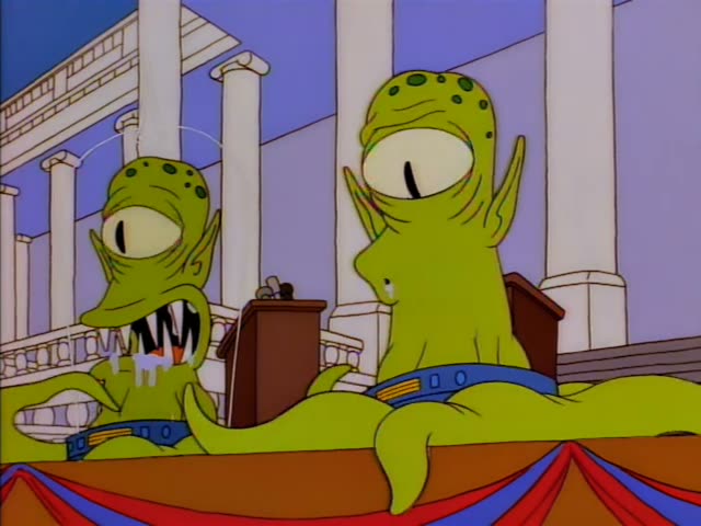 Two green, one-eyed aliens with spotted heads, seated behind wooden podiums on a columned stage draped in red, white…